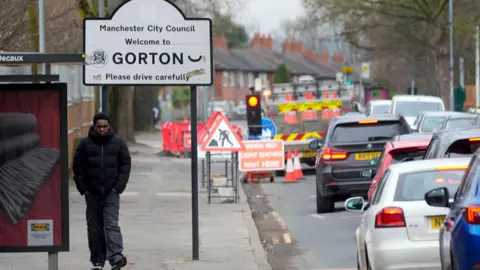 PA Media Generic street shot featuring a sign denoting the district of Gorton. There is a traffic jam and a teenager wearing a black puffa jacket, jeans and trainers walking along the pavement. 
