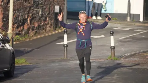 BBC A man with silver hair running on a road with his hands in the air. He has a mobile phone in his hand and a pair of white-framed sunglasses on his head.