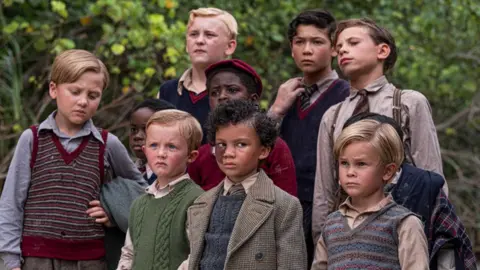 BBC/Eleven/J Redza A group of young actors stand together while filming Lord of the Flies. They are dressed in 1950s clothing and many of the boys have looks of concern on their faces.