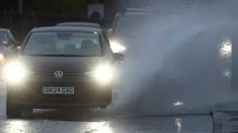 Reuters A small black Volkswagen car with its headlights on drives through a large puddle and throws up a tall stream of water onto the pavement.