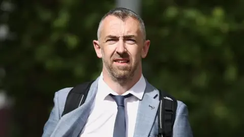 PA Media Daithí McKay is wearing a white shirt and a navy tie. He has a grey blazer on and a black rucksack. He has short grey hair and a beard.