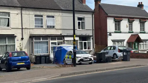 A street, where one of the semi-detached houses is taped off with police tape. A police car and blue West Midlands Police tent are positioned outside one of the houses.