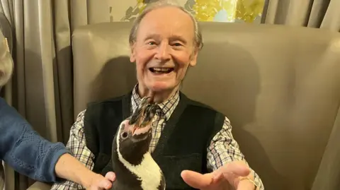 A resident at the care home is holding a penguin on his lap. He is smiling and pointing at the camera. 