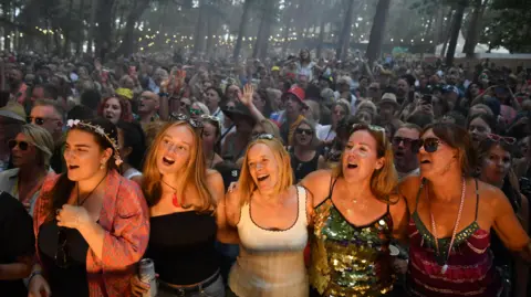 Five women at the front of a packed crowd. They are illuminated by lights from the stage and they have got their arms around each other as they sing along to an artist, who cannot be seen. 