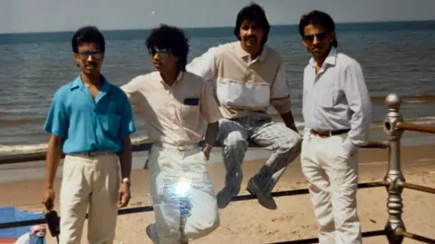 Family photograph Masrob Ali, in blue, pictured a few years after arriving in the UK from Bangladesh, alongside three other men sitting across a fence at a beach
