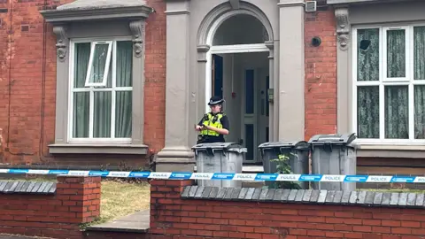 BBC A police officer in uniform and high-vis vest stands in front of a brick building. There is blue striped police tape in front of a building in line with a low wall