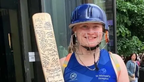 Isla Bayliss is wearing cricket gear, including a blue helmet, and holding a cricket bat with extensive writing on it.