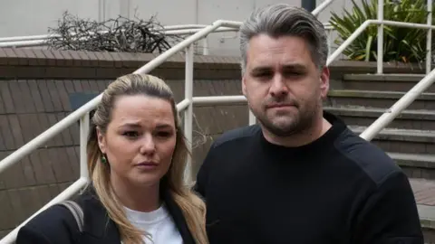 John Fairhall/BBC Bryony and Daniel are standing outside the coroner's court, in front of the steps, both wearing black. Bryony is shorter and has blonde hair. Daniel has grey hair and is unshaven.