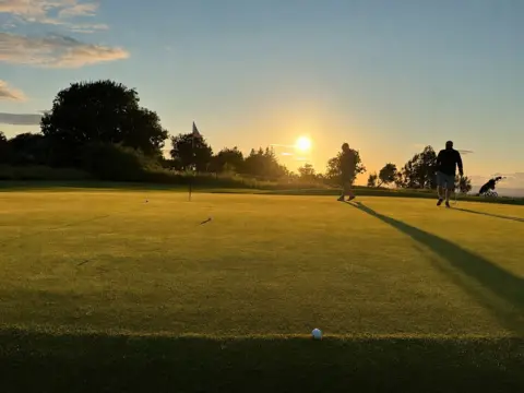 Thomas Martin The sun setting over a golf course, with two men still out on the grass, walking towards the hole while carrying their clubs.