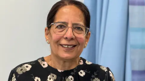 A woman with long dark hair and a dark patterned top. She is smiling at the camera while sitting in a hospital ward. A blurred blue curtain can be seen behind her.