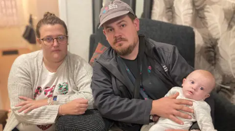 A woman with glasses in a grey cardigan sitting next to a man in a black jacket and grey cap. He is sitting on a black chair and is holding a baby