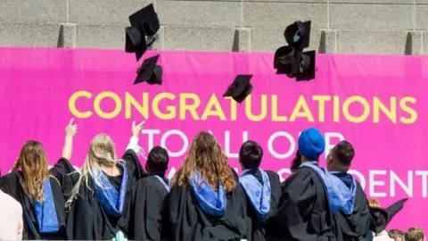 University of Brighton Graduation celebrations are taking place in Brighton and students are standing in front of a pink congratulations sign. They have their backs turned so the hoods on their academic gowns are visible. They have thrown their mortarboards into the air.