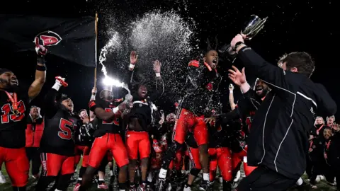 @grapplingwithimages A group shot of the Bullets at night on the pitch celebrating, spraying champagne in the air. They are in red and black American football kits and are jumpingin the air while someone in front of them holds a trophy.