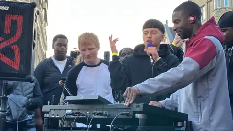 Three men stand in front of a DJ mixer with people gathered behind them. Ed Shearan, on the left, has short ginger hair and is wearing a white t-shirt with black trim markings. Next to him is a young man with a microphone in his left hand and on the far right is a man wearing a grey and red hoody with his hands on the mixing table.