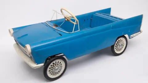 A child's pedal car dating from the 1950s. It is blue, with a white steering wheel and front bumper, and is in the typical boxy shape of the time.