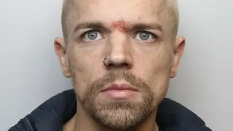 A man with facial hair, a nose piercing and a graze at the op of his nose, wearing a black jacket. It is a custody photo.