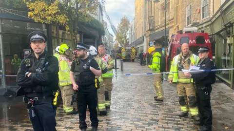 Simon Jones/BBC Police office and firefighters standing by blue and white police tape with fire vehicles and a ladder in the background.