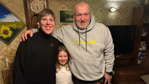 Mark Russo stands on the right in a grey hoodie with his arm around his wife. His daughter stands in the middle, a little girl in a white shirt, Mrs Russo stands wearing a black jumper on the left.