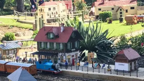 Miniature model railway station with trains on tracks in front and model people on a platform.