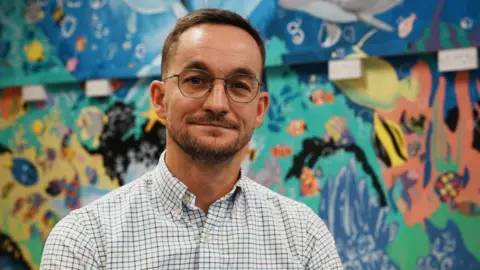 John Fairhall/BBC Tom Abell sits in front of a colourful wall mural. He is wearing a checked shirt, spectacles and has a short beard.