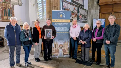 Contributed A group of people associated with St Mary's Church stand together in a group and smile at the camera. In the middle of them is a tall information board about their heating fund. One of the men, the Reverend Nigel Prior, holds the Maggi Hambling painting in his hands. 
