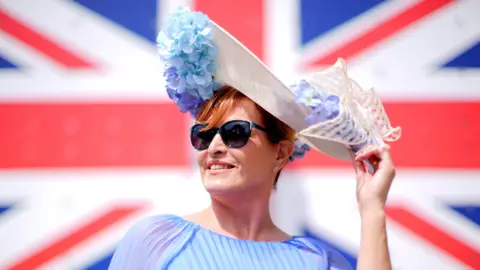 A woman with a floral hat standing in front of a Union flag. She is also wearing a light blue dress and sunglasses.
