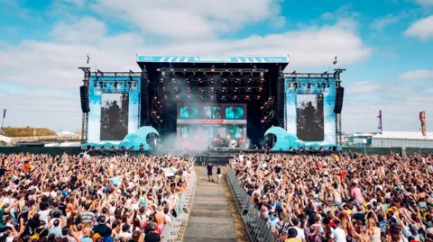 Will Bailey A huge crowd stretches out in front of a large outdoor stage at Boardmasters, with festivalgoers raising their hands as a performance takes place under bright daylight.