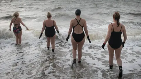 PA Media This image shows four people in swimming costumes entering the sea. Some are also wearing wetsuit boots. The water appears to be fairly rough, with waves lapping the beach which appears to be shingle or sand. All four people have their backs to the camera.
