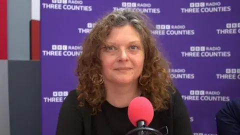 Close-up of Zara Layne seated in a radio studio, positioned behind a microphone with a red foam cover. The background features a purple wall repeating the text “BBC Radio Three Counties.” The person has curly medium-length hair and is wearing a dark top, with studio equipment visible in the foreground.