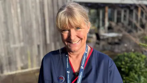 Juliette Hughes wearing dark blue hospice scrubs and a branded lanyard. She has blonde hair which is tied back into ponytail and styles with a fringe. She is standing outdoors on a bright sunny day in front of a wooden fence and some bushes, smiling at the camera.