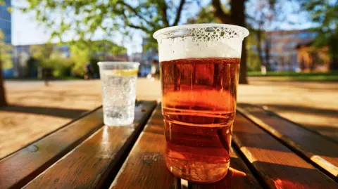 Plastic cups of beer and water sit on an outside wooden table. The background is blurred  but contains the outline of green trees and blue sky. 