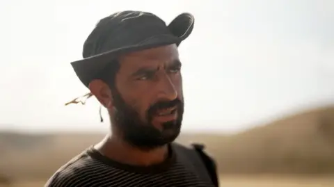 Ahmad Daraghme is photographed at shoulder height with the background blurred. He wears a hat and a black beard and looks off-camera to his left.