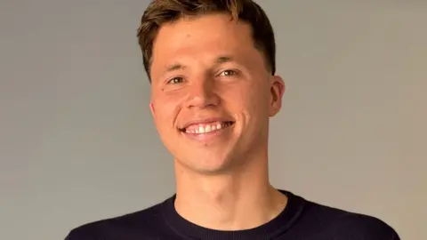 Freddie Woodward: a young man with short dark hair sits on a grey sofa, smiling at the camera. He has short brown hair and wears a navy jumper.