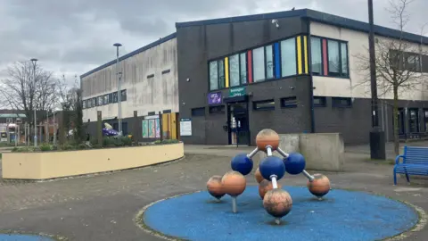 Photograph of Brinnington Library in Stockport. The image shows a sculpture and planters in front of the area's shopping precinct.