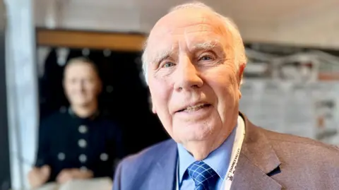Head and shoulder photo of Ken Wells who has short grey receding hair and is smiling to camera. He's wearing a blue shirt suit and tie. The interior of the police museum is seen in the background out of focus, including the mannequin of a victorian police officer.