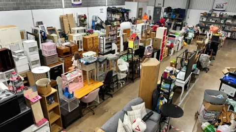 A warehouse full of items including furniture, toys, books and decorations. They are arranged in rows like aisles for people to walk between