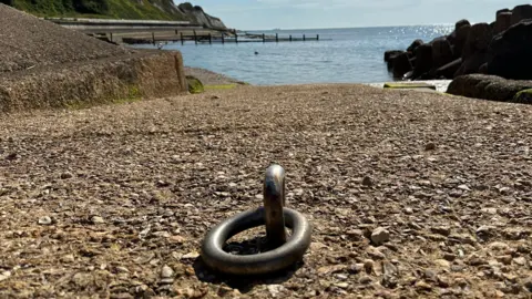 Daisybell A concrete slipway slopes towards the sea with a round metal mooring ring dominating the foreground. A calm blue sea can be seen in the background with rocks to the right and wooden breakwaters to the left.