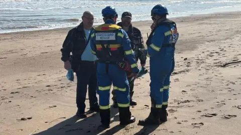 Kevin Shoesmith/BBC Stuart Green dan pria lain berbicara dengan pejabat CoastGuard, yang mengenakan seragam dan helm biru, di pantai yang cerah. 