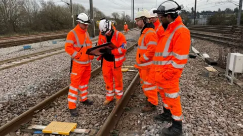 Five men in hi-viz outfits and helmets stand on a rail track. One is looking at a plan while the others look on.