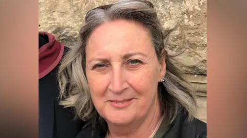 A woman with short grey hair wearing a black jacket, khaki green shirt, and a bag with a gold chain. She is smiling at the camera and has a pair of sunglasses on her head.