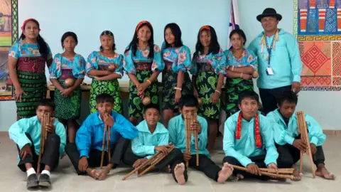 Seven girls are standing behind six boys with their teacher. Boys are wearing turquoise shirts, the girls are wearing black and green moles wrapped over the waist and turquoise floral blouse. The backgrounds have bright colored wall hanging.