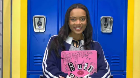 Vivian Panka stands against a blue and yellow locker on stage wearing a blue jacket with white stripes and a chequered blue and black dress. She holds a pink Burn Book and has long black hair
