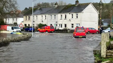 Red vehicles parked at the top of a hill as flood water runs down the road. One of the vehicles, to the right, is moving through the flood water. 