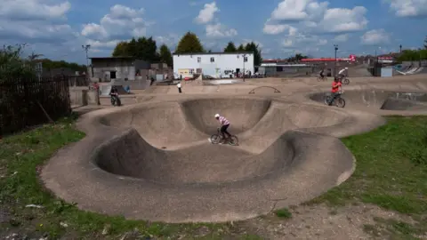 Getty Images The Rom a 1970s skate park made of what appears to be brown coloured concrete is being used by several young people on bikes. 