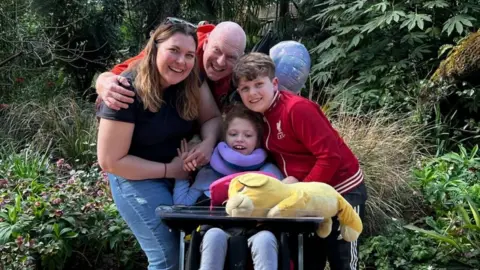 Addy poses for a family photo with her mother, father and brother in front of plants and flowers. She is in a specialist wheelchair. 