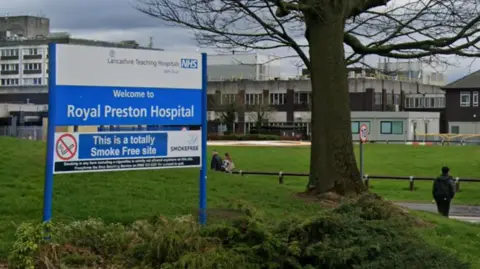 A blue-and-white sign saying "Welcome to Royal Preston Hospital" in lawned gardens at the entrance. There are buildings in the background, a large tree to the right and people milling about in the grounds.