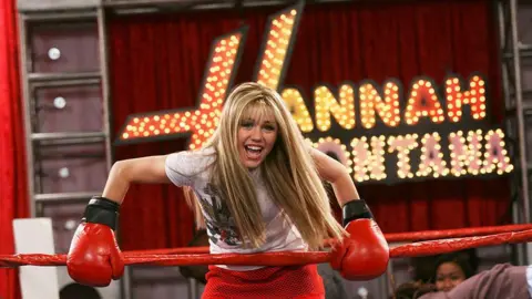 Getty Miley Cyrus is shown as her popstar alter ego Hannah Montana. She wears red boxing gloves and is stood in a ring with the Hannah Montana sign behind her