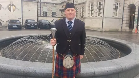 Jason Price is wearing a red and navy tartan kilt and sporran and is holding a mace. He is also wearing a navy jacket, striped tie and navy hat
