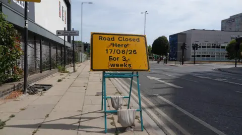A yellow sign on the pavement says " Road Closed Here 17/08/26 for 3 weeks".  The sign is weighted down with sand bags.  The road behind it has two lanes with arrows pointing in a direction away from the sign.