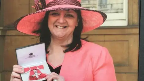 Supplied Wendy is wearing a pink hat and jacket. She is holding her MBE